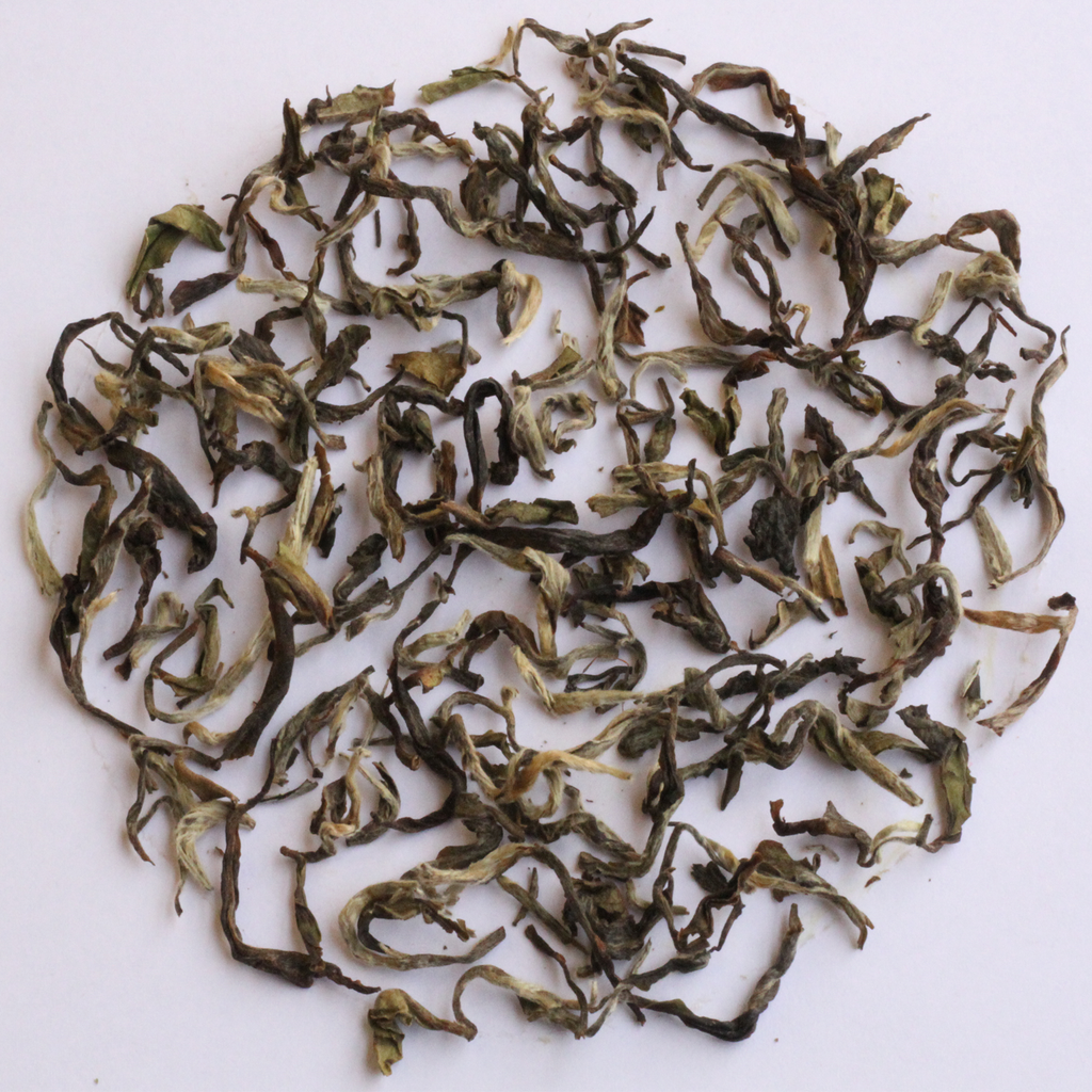 Loose-leaf tea leaves, characterized by their curly, twisted shapes and mix of dark green, olive, and light yellowish-green tones, arranged in a circular pile on a plain white background.