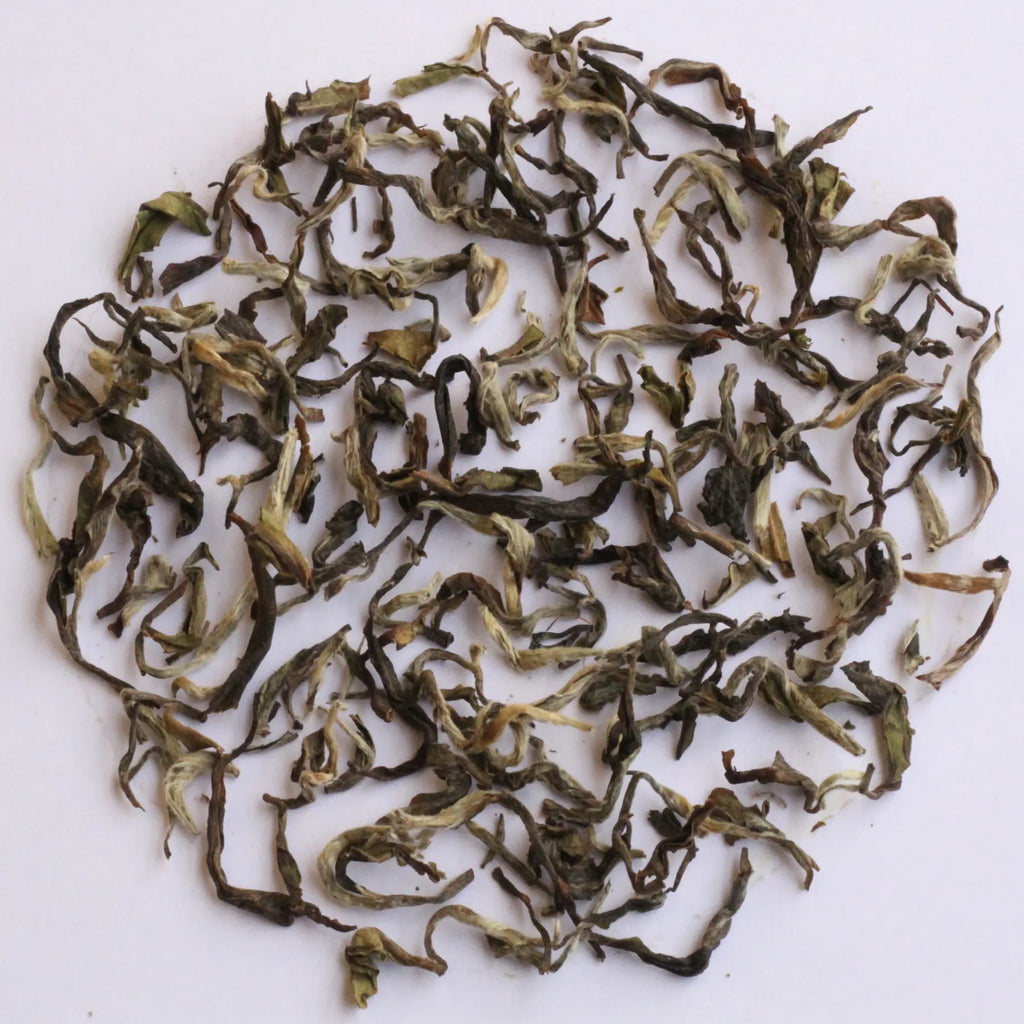 Loose-leaf tea leaves, characterized by their curly, twisted shapes and mix of dark green, olive, and light yellowish-green tones, arranged in a circular pile on a plain white background.