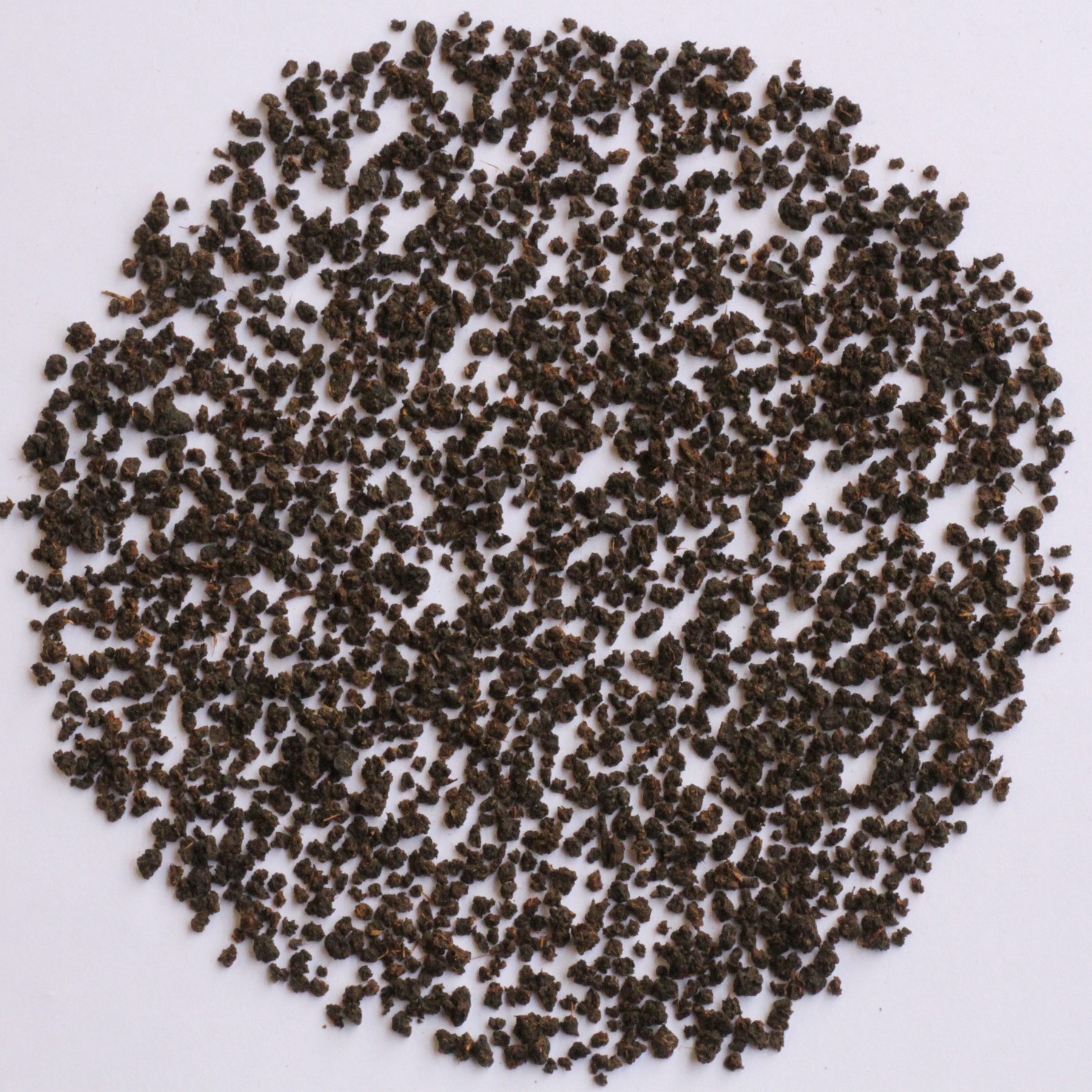 A pile of small dark brown to black granular particles or seeds arranged in a circular shape on a plain white background.