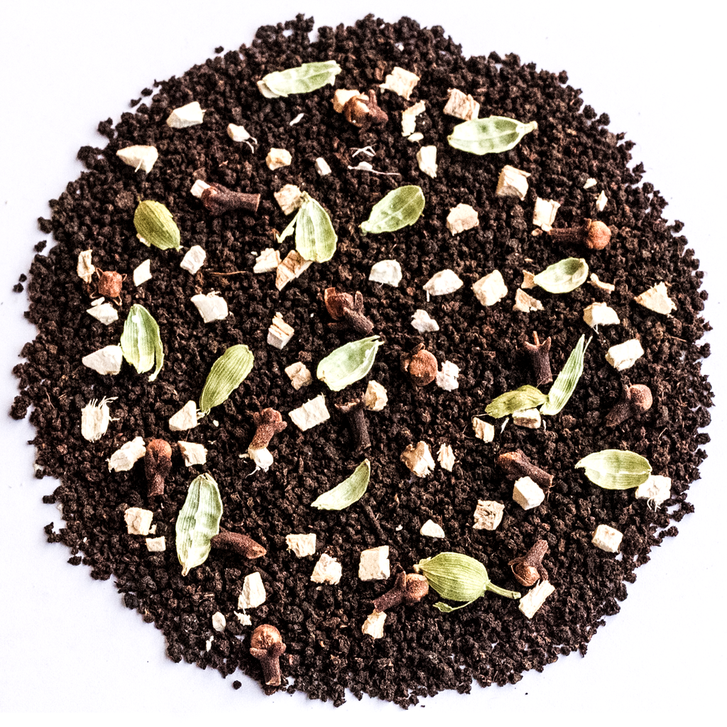 A circular pile of dark chocolate-flavored coffee mix or cocoa powder, featuring whole coffee beans, green cardamom pods, cinnamon sticks, and small white chunks, photographed from directly above on a white background.