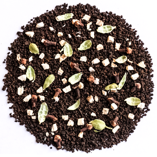 A circular pile of dark chocolate-flavored coffee mix or cocoa powder, featuring whole coffee beans, green cardamom pods, cinnamon sticks, and small white chunks, photographed from directly above on a white background.