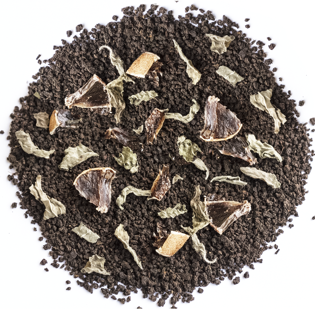 A top-down view of a circular pile of dark brown coffee grounds mixed with dried herbs, leaves, and small brown fruit pieces on a plain white background.