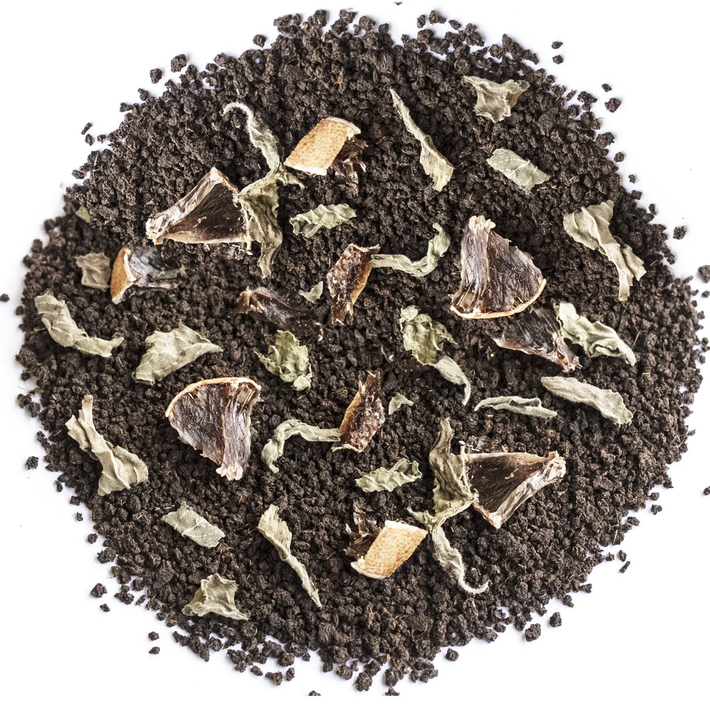 A top-down view of a circular pile of dark brown coffee grounds mixed with dried herbs, leaves, and small brown fruit pieces on a plain white background.