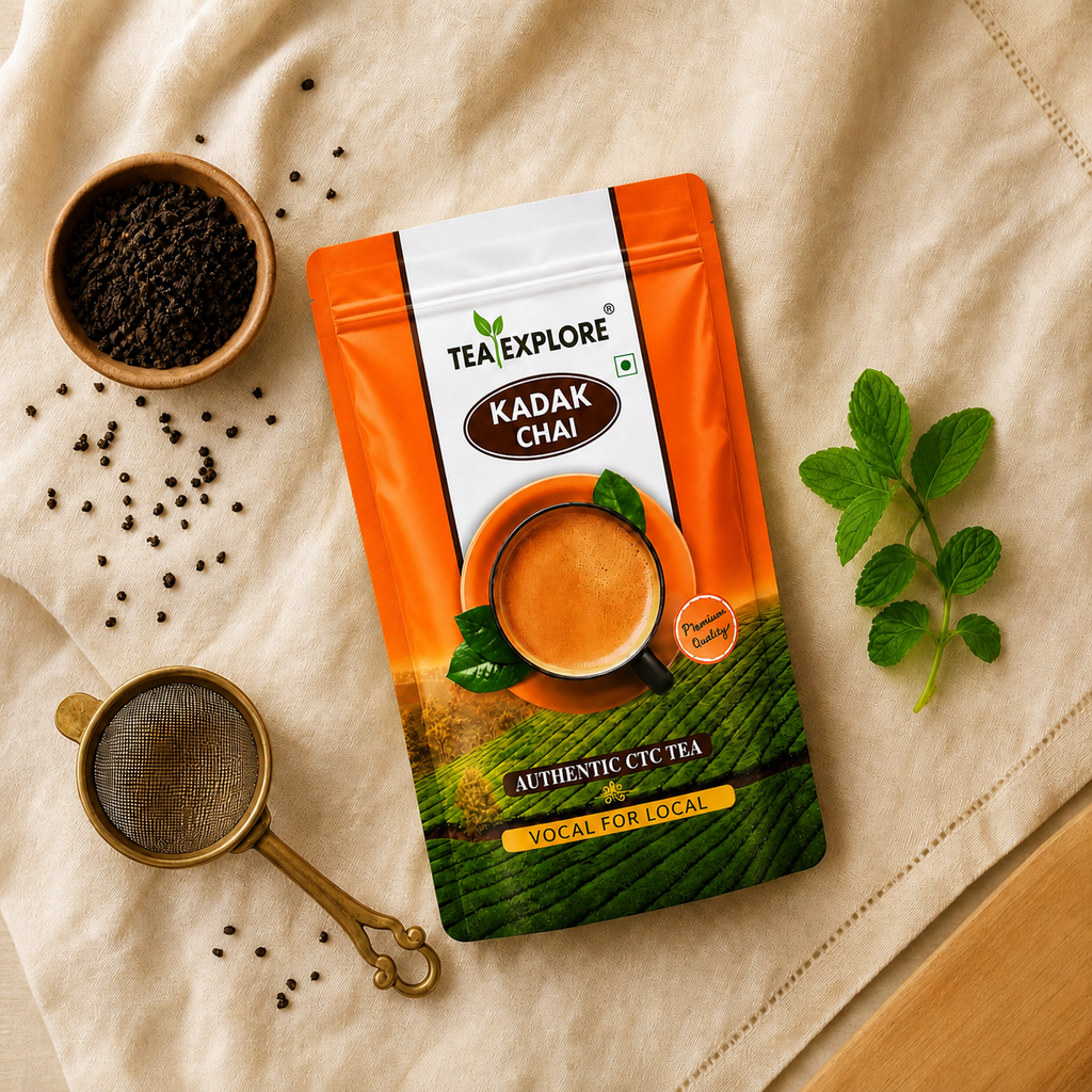 A package of Tea Explore Kadak Chai tea is displayed in a styled flat-lay arrangement on a beige fabric background, surrounded by whole black peppercorns in a wooden bowl, a brass strainer, and fresh mint leaves.