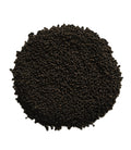 A pile of small dark brown to black granular seeds or pellets, photographed from above on a plain white background.